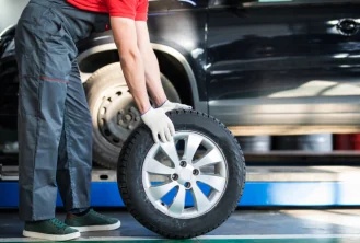A technician rolling a mounted wheel across a workshop floor toward a car.